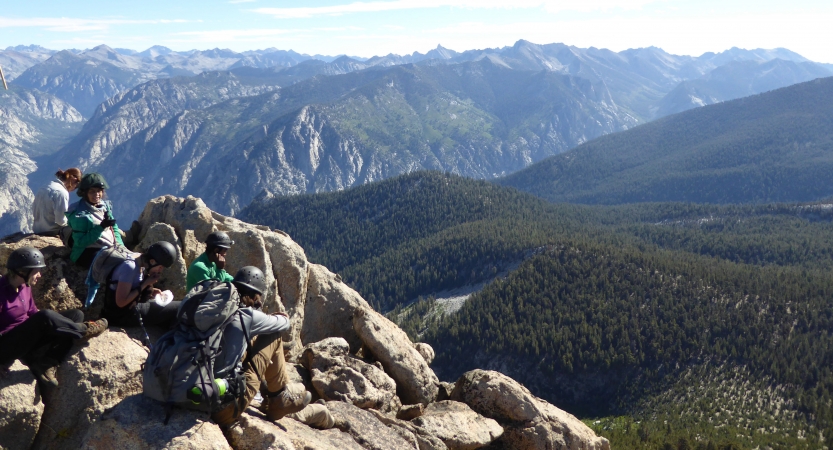 A group of people wearing helmets sit on rocky overlook, taking in the mountain landscape below. 